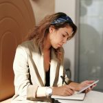 Confident businesswoman writing notes while using a smartphone in a sunny, modern office.